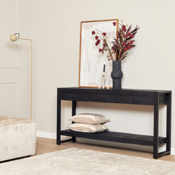 De Sidetable Boaz Zwart | 150 cm staat tegen een beige muur, gestyled met een vaas gedroogde bloemen, een ingelijste foto en glasdecor. Op de onderste plank liggen twee decoratieve kussens. Vlakbij staan een beige bank en een staande lamp op een licht vloerkleed.