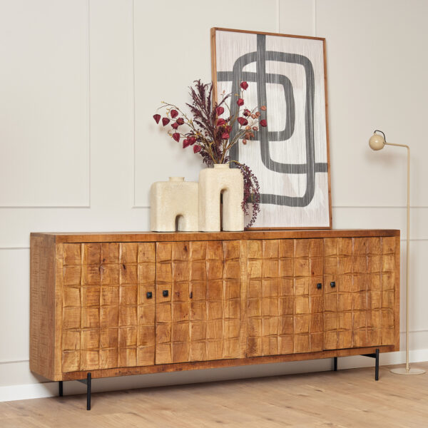 De Dressoir Brandy 220 cm, met een gestructureerde voorkant, staat tegen een lichte muur. Bovenop staan twee crèmekleurige vazen met gedroogde bloemen en een ingelijst abstract kunstwerk, terwijl rechts een moderne staande lamp staat.