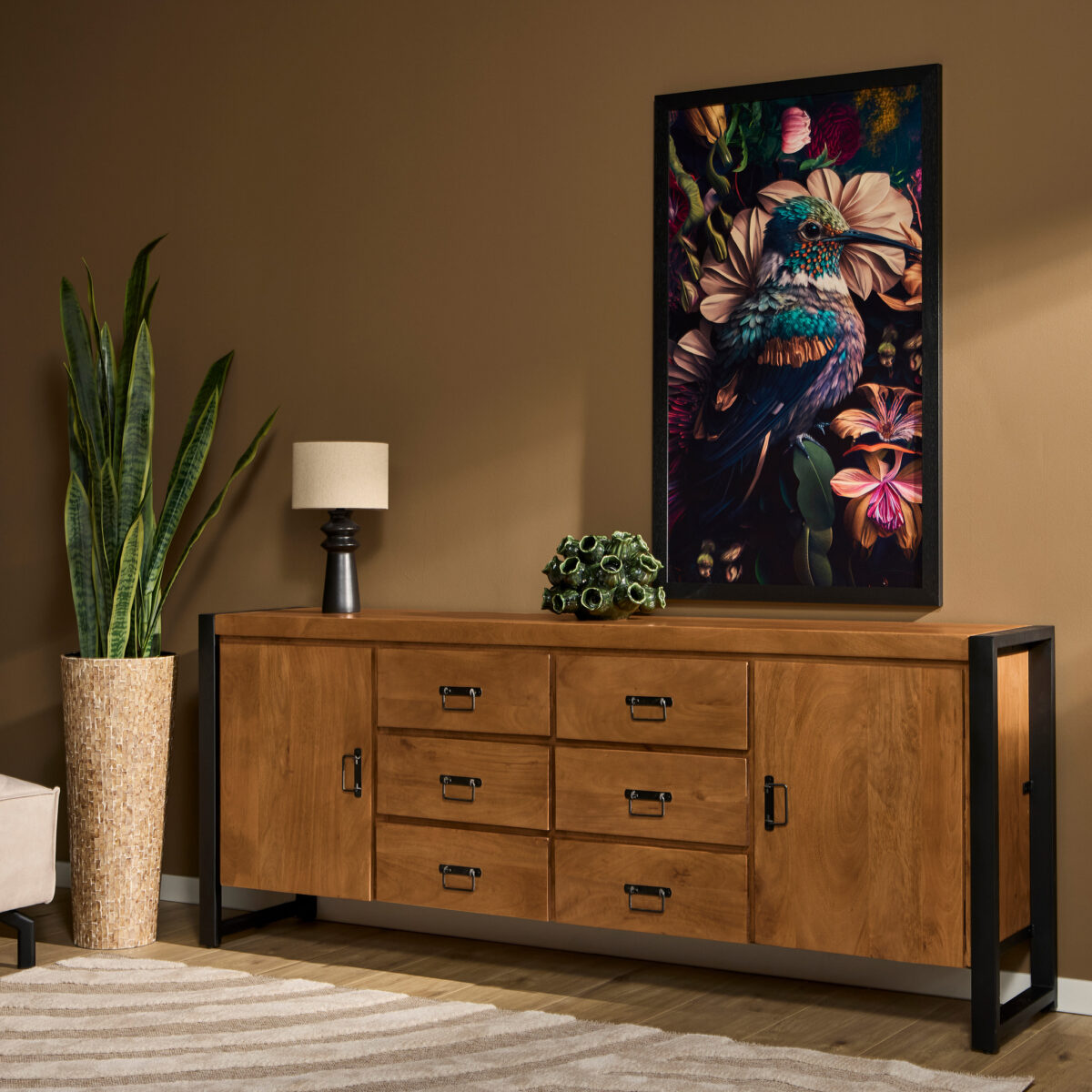 Een moderne kamer met het Dressoir Boaz 200 cm houten dressoir, een slangenplant in pot, een kleine lamp, een decoratief object en kleurrijke vogelkunstwerken op de bruine muur erboven. Een beige vloerkleed is gedeeltelijk zichtbaar op de vloer.