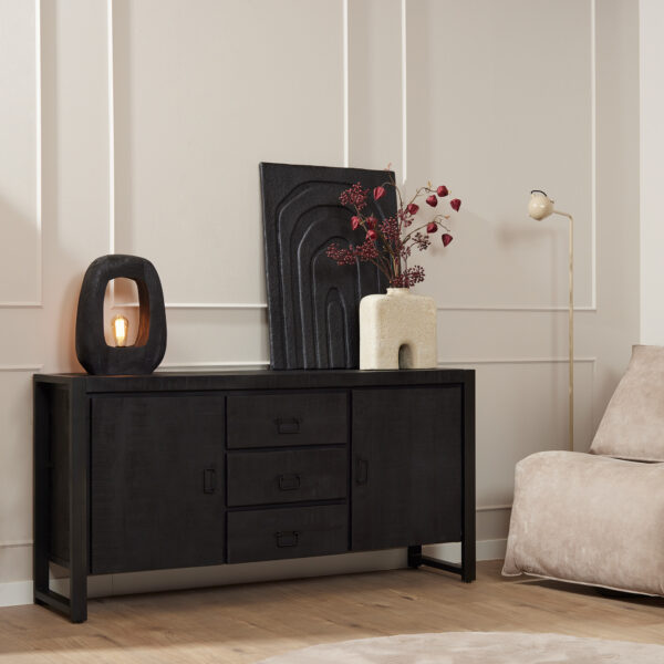 Een moderne woonkamer met de Dressoir Boaz Black (160 cm), zwarte muurkunst, een vaas met rood gebladerte, een sculpturale lamp, beige fauteuil en staande lamp tegen muren met crèmekleurige panelen en een houten vloer.