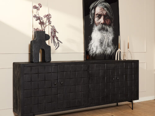 De Dressoir Brandy Black 220 cm staat tegen een lichte muur met daarop een sculpturale vaas met paars gebladerte, twee kandelaars en een ingelijst portret van een oudere man met een lange witte baard en hoofddoek.