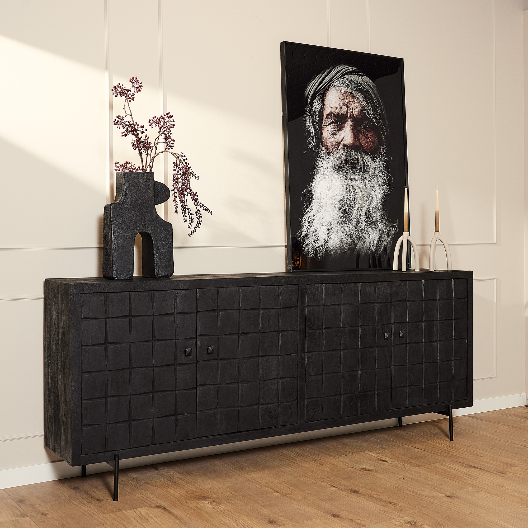 De Dressoir Brandy Black 220 cm staat tegen een lichte muur met daarop een sculpturale vaas met paars gebladerte, twee kandelaars en een ingelijst portret van een oudere man met een lange witte baard en hoofddoek.