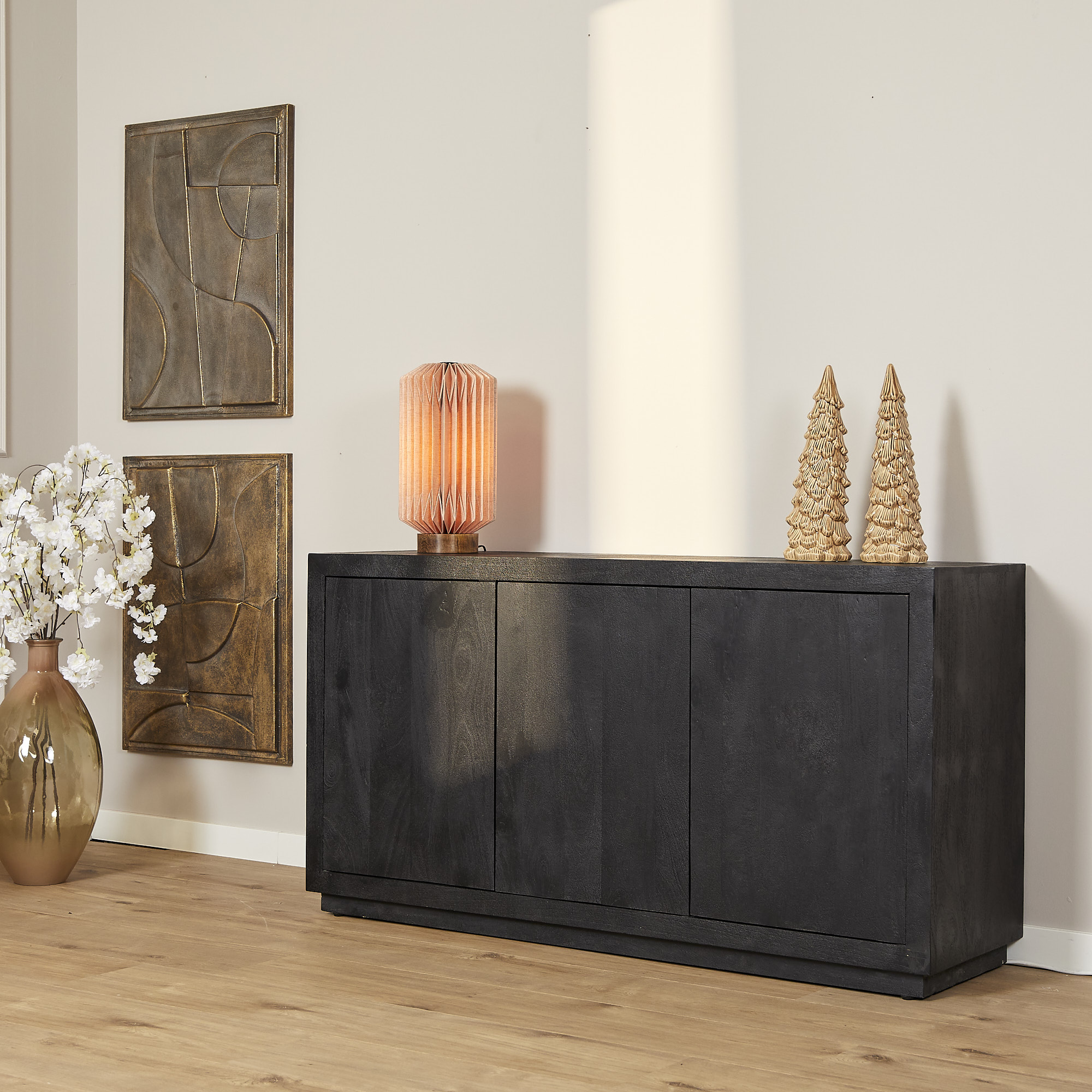 De Dressoir Brussel Black 3 deurs (150 cm) staat tegen een beige muur, versierd met een gloeiende oranje lamp, twee gouden kegelvormige ornamenten, een grote witte bloemenvaas en aangevuld met abstracte muurkunst.