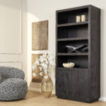 Een moderne woonkamer met de Boekenkast Brussel Zwart 90 cm met decor, een fauteuil met lichte structuur, een grote gouden vaas met witte bloemen en twee abstracte gouden muurkunstwerken op beige muren en een lichthouten vloer.