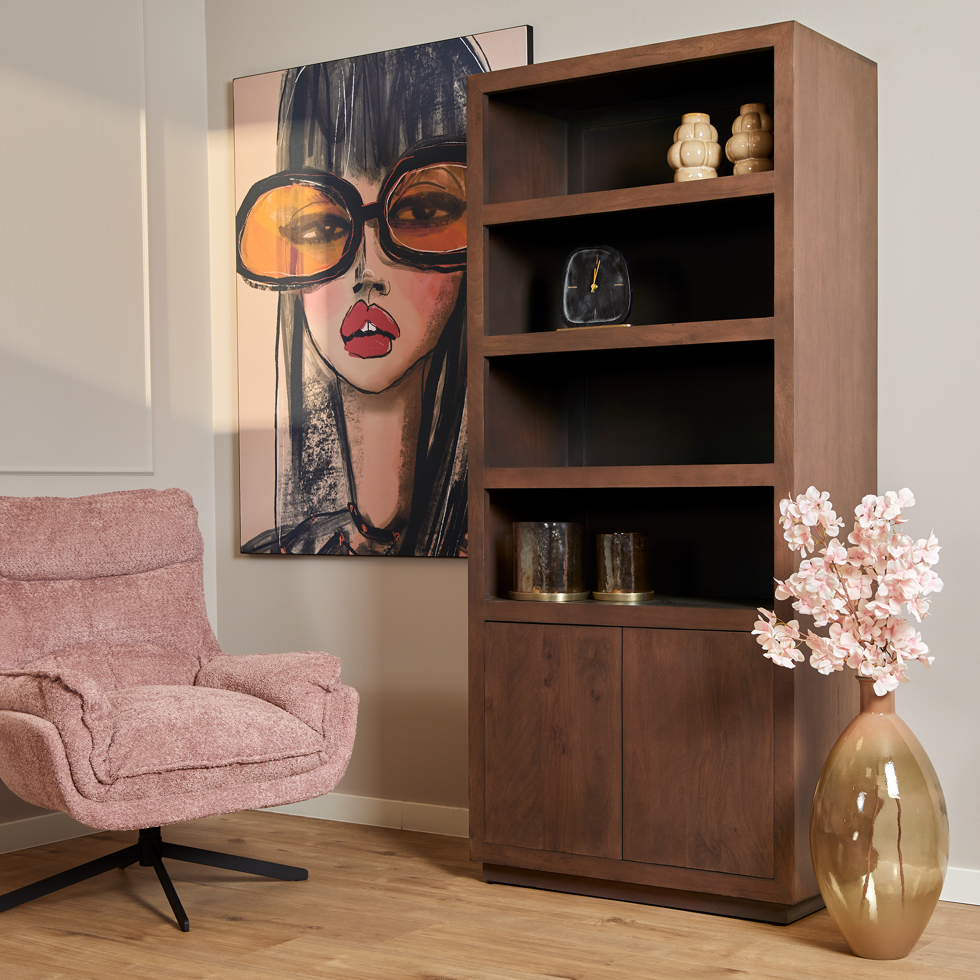 Een moderne woonkamer is voorzien van een roze fauteuil met structuur, de Boekenkast Brussel Walnut (90 cm) met decoratieve items, een grote vaas met roze bloemen en muurschilderingen van een vrouw met een grote bril.