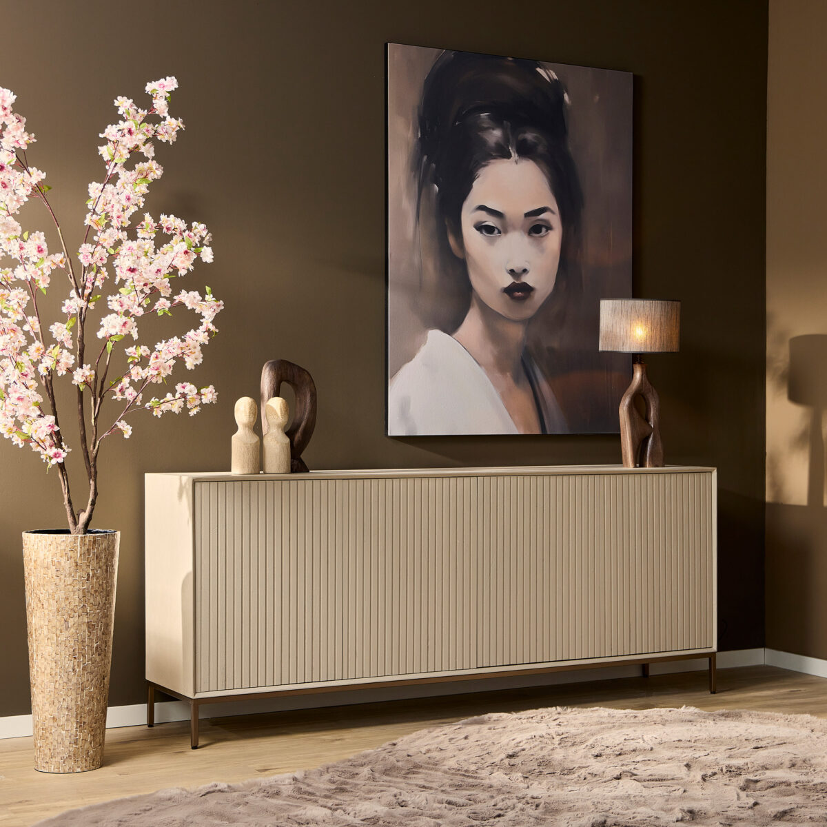 In een moderne kamer staat de Dressoir Madison Sand | 210 cm onder een groot vrouwenportret, in combinatie met een sculpturale tafellamp, abstract decor en een hoge vaas met roze bloesemtakken tegen een bruine muur.