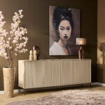 In een moderne kamer staat de Dressoir Madison Sand | 210 cm onder een groot vrouwenportret, in combinatie met een sculpturale tafellamp, abstract decor en een hoge vaas met roze bloesemtakken tegen een bruine muur.