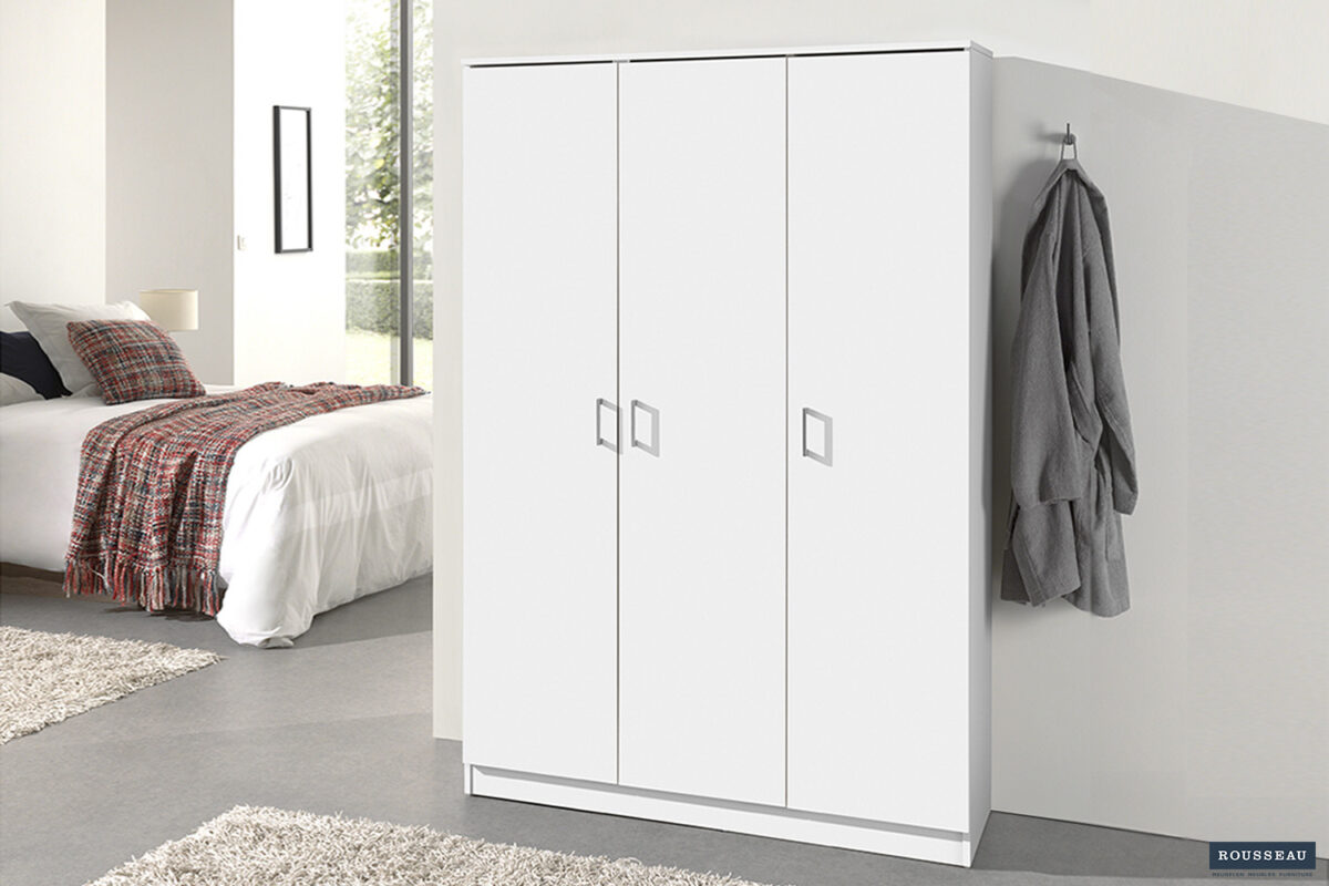 De Opbergkast 'Ray' 3-deuren en 4 legplanken in wit staat in een moderne slaapkamer naast een muur met een grijze jas aan een haak. Op de achtergrond zie je een bed met plaid dekentje en een raam.