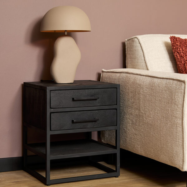 Een modern Nachtkastje New York met twee lades bevat een sculpturale beige lamp. Ernaast staat een crèmekleurige bank met een rood kussen, tegen een mauve muur en houten vloer.