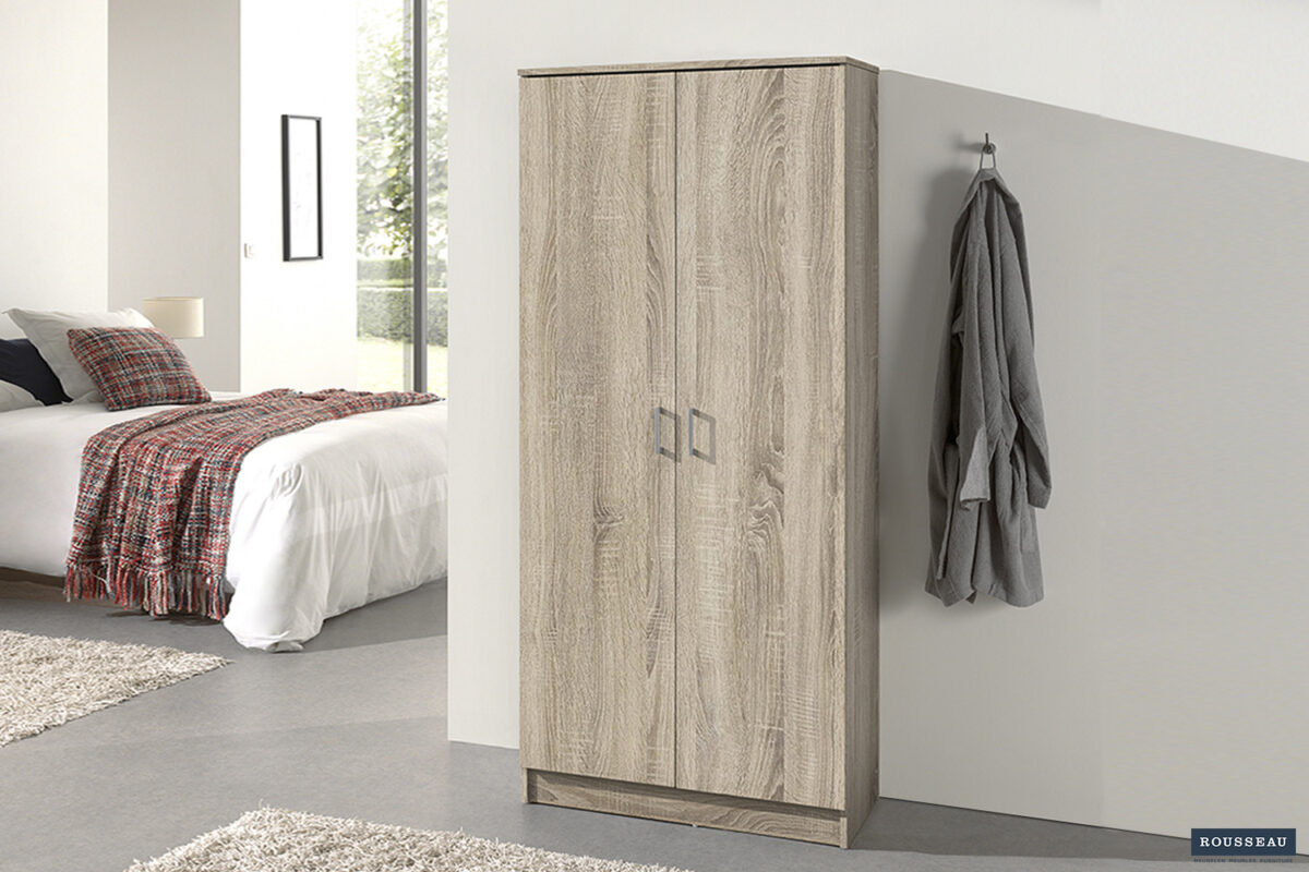 Een moderne slaapkamer met de Opbergkast 'Ray' 2 deuren en 4 legplanken Sonoma, een wit bed met een geruite deken, twee beige vloerkleden en een grijze badjas aan een wandhaak. Zonlicht stroomt naar binnen vanuit het raam op de achtergrond.