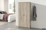 Een moderne slaapkamer met de Opbergkast 'Ray' 2 deuren en 4 legplanken Sonoma, een wit bed met een geruite deken, twee beige vloerkleden en een grijze badjas aan een wandhaak. Zonlicht stroomt naar binnen vanuit het raam op de achtergrond.