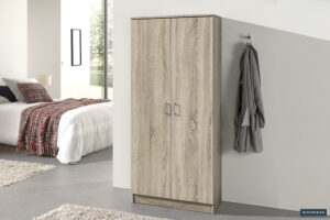 Een moderne slaapkamer met de Opbergkast 'Ray' 2 deuren en 4 legplanken Sonoma, een wit bed met een geruite deken, twee beige vloerkleden en een grijze badjas aan een wandhaak. Zonlicht stroomt naar binnen vanuit het raam op de achtergrond.