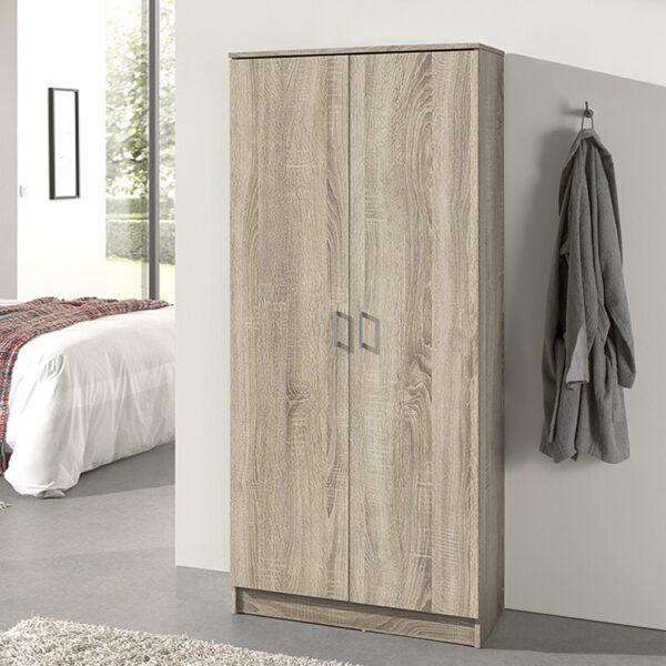 Een moderne slaapkamer met de Opbergkast 'Ray' 2 deuren en 4 legplanken Sonoma, een wit bed met een geruite deken, twee beige vloerkleden en een grijze badjas aan een wandhaak. Zonlicht stroomt naar binnen vanuit het raam op de achtergrond.
