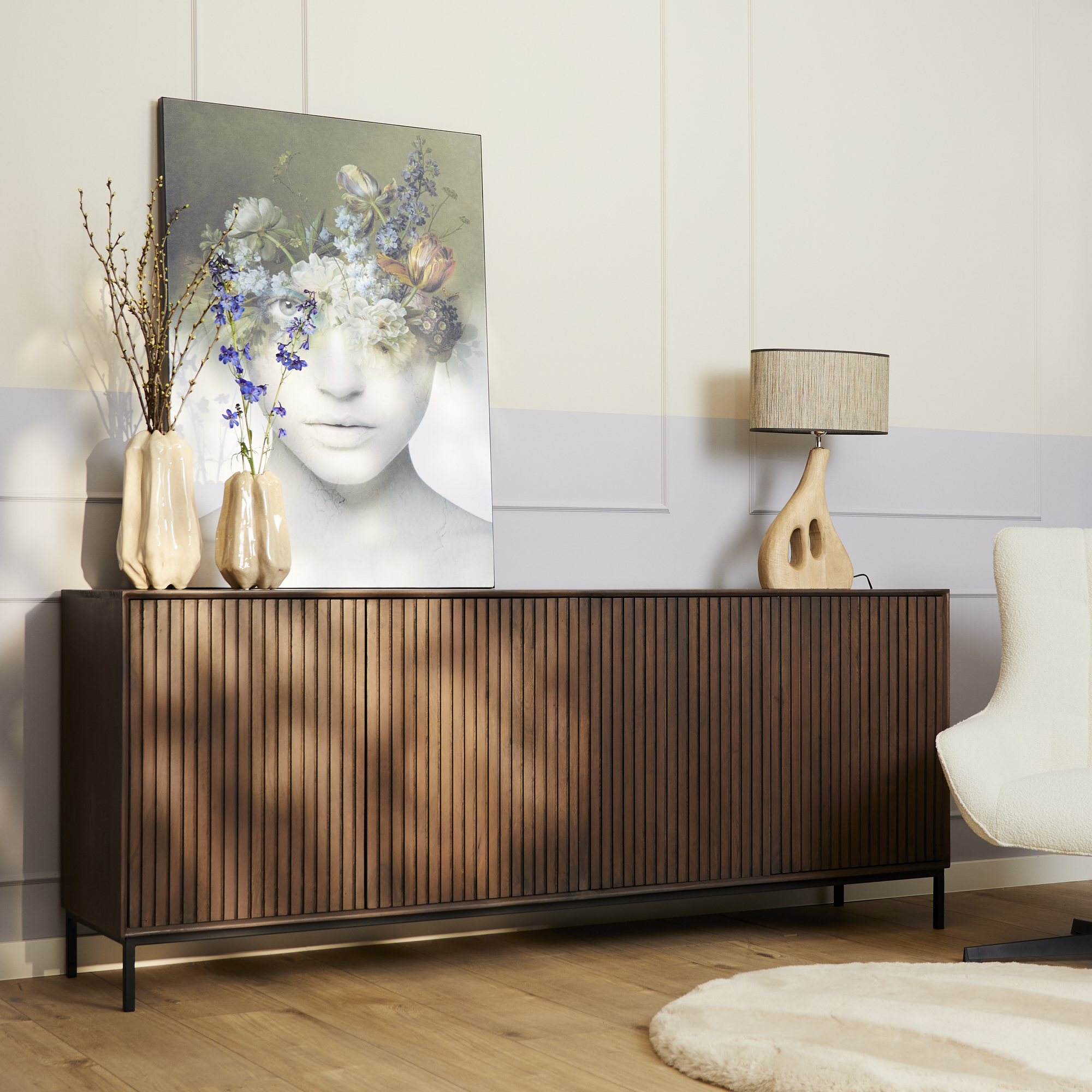 Een moderne woonkamer met de Dressoir Madison Walnoot (210 cm), decoratieve vazen met bloemen, een tafellamp, abstracte bloemenportretkunst, een witte stoel en een rond beige vloerkleed op een houten vloer.