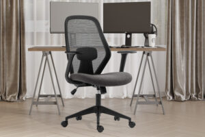 De Bureaustoel 'Claude' Mesh Zwart/Grijs (1c), een moderne ergonomische stoel met netrugleuning en verstelbare hoofdsteun, staat voor een houten bureau met twee monitoren, toetsenbord, muis en beker in een thuiskantoor met beige gordijnen en houten vloeren.