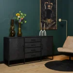 Een moderne woonkamer heeft een donkergroene muur, Dressoir New York | 165 cm in zwart, vazen met bloemen, een ingelijst portret, twee witte kaarsen, een tan fauteuil en een rond zwart vloerkleed op een lichte houten vloer.