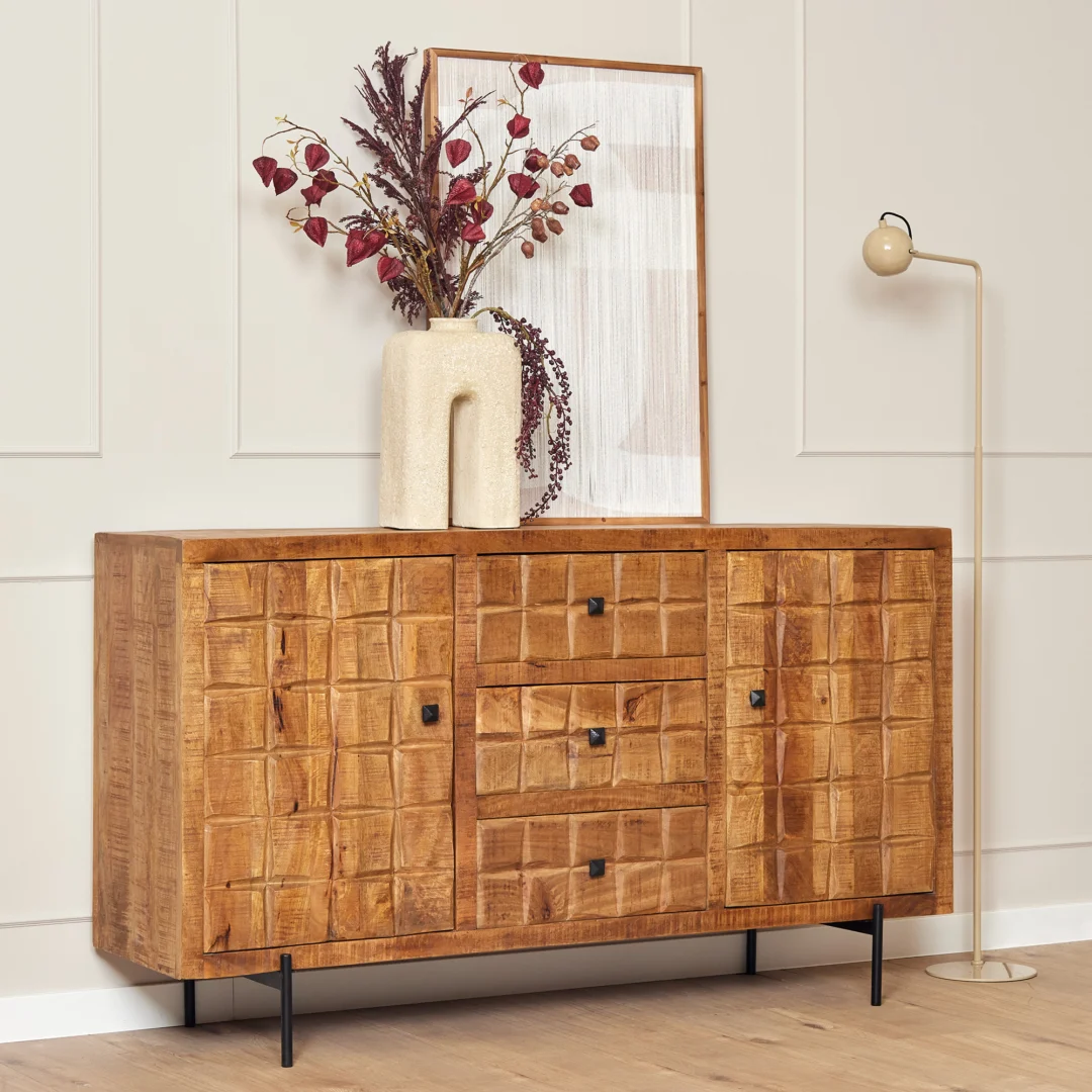 De Dressoir Brandy | 160 cm heeft een geometrische houten voorkant en staat tegen een crèmekleurige muur. Het is vormgegeven met een sculpturale vaas met gedroogde bloemen, ingelijst kunstwerk bovenop en wordt aangevuld met een beige vloerlamp.