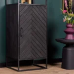 De Boekenkast New York, gemaakt van mangohout en staal met een zwarte deur met chevronpatroon, staat op een houten vloer naast een ronde zwarte tafel met een paarse vaas met kleurrijke bloemen tegen een donkergroene muur.