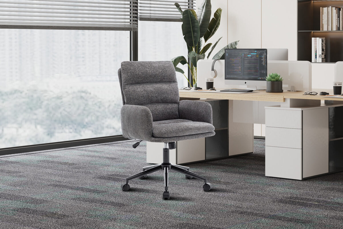 In een modern kantoor met een groot raam staat de Bureaustoel 'Aimé' Stof Fabb Raven 13 Donkergrijs, een strakke grijze bureaustoel op wieltjes, naast een houten bureau met een computer, laden en een plant in pot met uitzicht op gebouwen in de stad.