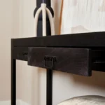 Een close-up van de Sidetable Boaz Black 120 cm toont het moderne zwarte houten ontwerp met een open lade en metalen handvat, aangevuld met decoratieve items zoals een zwart-witte vaas en ingelijst kunstwerk.