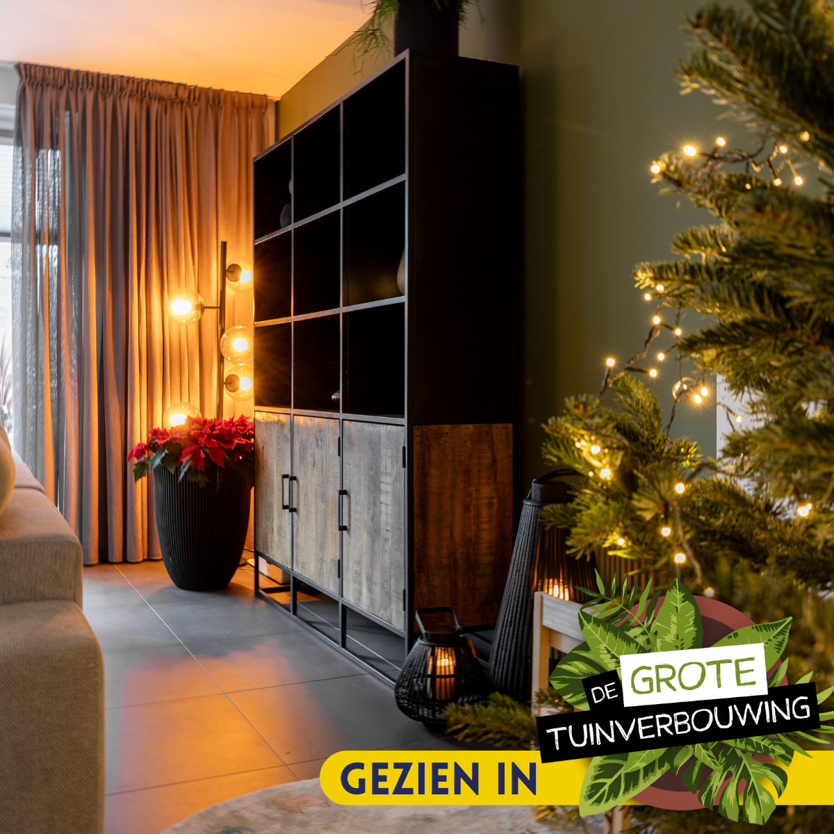 Een gezellige woonkamer met de Boekenkast Oakland XXL 160 cm, een hoge plant, warme vloerverlichting, beige gordijnen en een versierde kerstboom op de voorgrond. Het "Gezien in De Grote Tuinverbouwing" logo verschijnt in de hoek.
