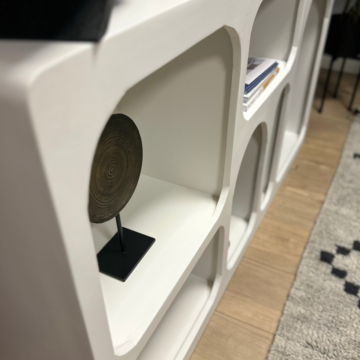 Een close-up van de Dressoir Caya Wit toont het moderne, witte ontwerp met afgeronde rechthoekige vakken. Op één plank staat een rond decoratief stuk, terwijl op een andere plank gestapelde boeken staan. De houten vloer en een deel van een tapijt met patroon zijn eronder zichtbaar.