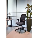 De Workliving Nora Mesh Zwart Regain - Bureaustoel (NEN1335 Ergonomisch Design) staat naast een bureau met een lamp en een koffiekopje, bij een grote potplant in een minimale, goed verlichte kantoorruimte.