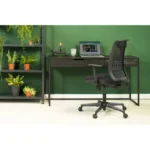 Een modern thuiskantoor met een zwart bureau, open laptop, kaars, pen en notitieblok. De Workliving Base Mesh - Ergonomische Bureaustoel (N)EN 1335 staat op het bureau. Groene muurachtergrond; links een plank met potplanten.