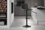 Een moderne keuken met grijze kastjes, gestapeld haardhout en een enkele 'Trevi' PU Zwart barkruk bij een wit aanrecht op een betonnen vloer.