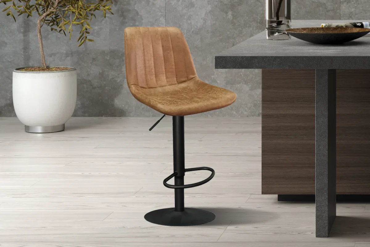 In een moderne keuken met een lichthouten vloer staat de 'Barcelona' barkruk in PU Ote Topper 02 Cognac met een zwart metalen onderstel naast een aanrecht van donker hout en grijze steen, terwijl een plant in pot op de achtergrond staat.