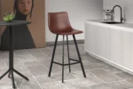 Een moderne keuken toont Barstoel 'Ozan' PU Bruin barkrukken met zwarte metalen poten, een ronde tafel met groene appels, een licht aanrecht met spoelbak en een groot vrouwenportret op de achtergrond.