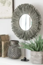 De Mirror On Foot Round Reed Raffia Grey, met zijn decoratieve stro-achtige frame en zwarte standaard, staat op een wit oppervlak naast een gevlochten metalen vaas, een potplant, boeken met patronen en een gedeeltelijk zichtbare wandkaart.