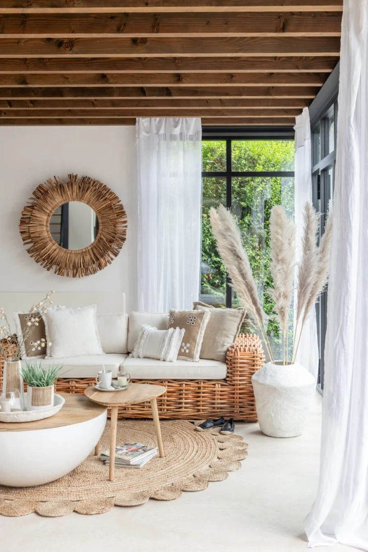 Een gezellige, zonovergoten woonkamer met een rieten bank, neutrale kussens, een rond geweven vloerkleed, een houten salontafel, pampagras in een witte vaas, transparante gordijnen en de spiegel Maurice Round Water Hyacinth Brown Large aan de muur.