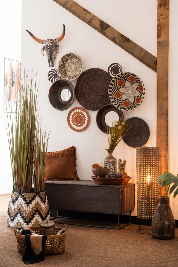 Een gezellige, moderne kamer met een bruine bank, decoratieve kussens, hoog gras in een mand, gevlochten manden, de spiegel Round Bowl Shaped Recycled Wood Brown aan de muur, Afrikaans geïnspireerde decoratie en warme verlichting van een staande lamp.