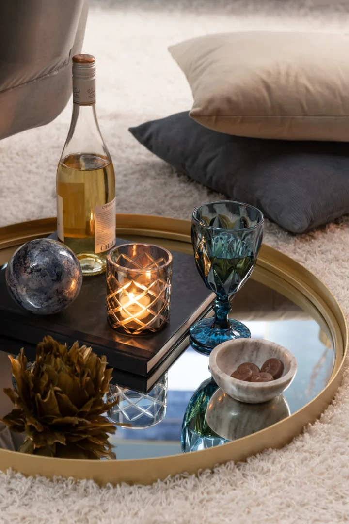 Op het kleine dienblad Mirror Round High Border Metal/Glass Gold staan witte wijn, een blauw glas, een brandende kaars, een decoratieve bol, chocolaatjes in een schaal en een goudkleurige bloem, met kussens op een pluche tapijt op de achtergrond.