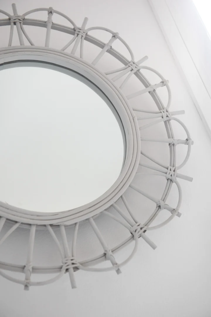 De spiegel Round Bamboo Light Grey, met een decoratieve geweven lijst, hangt aan een witte muur bij een raam en weerkaatst het natuurlijke licht.