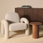 De Fauteuil Phiel - Bouclé Naturel staat naast een kleine ronde houten bijzettafel en een donkere houten kast met boeken, allemaal gerangschikt op een licht vloerkleed in een minimalistische kamer.