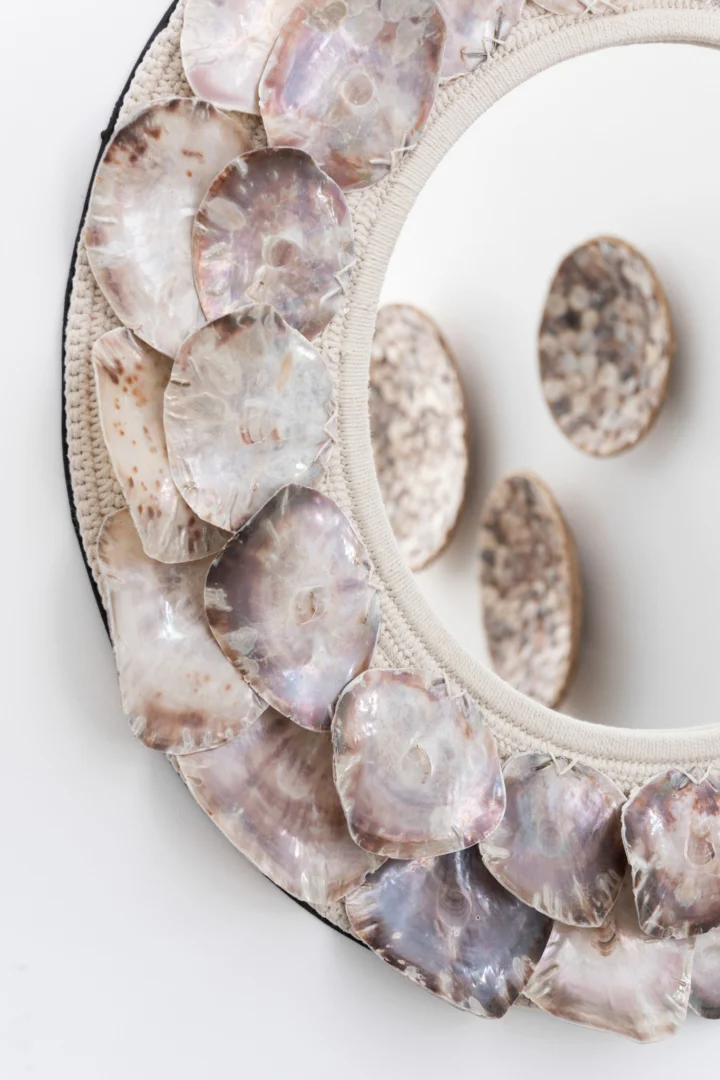 De Mirror Lisa Shells Purple Small is een ronde spiegel met een iriserende, met schelpen versierde lijst die meer met schelpen bedekte voorwerpen weerspiegelt tegen een witte achtergrond.