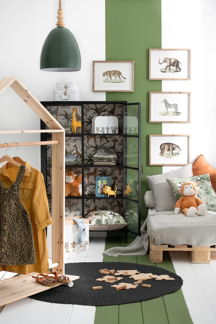 Kinderkamer met dierenthema, met Vloerkleed Rond Jute Zwart, ingelijste dierenprints aan de muur, een glazen kast met dierenfiguren, een pluchen aap op een palletbank, een houten kledingrek en groene accenten.