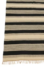 Het Vloerkleed Strepen/Stippen + Franjes Jute Ecru/Zwart is een geweven vloerkleed met afwisselend zwarte en beige strepen, gevarieerde patronen en texturen, en beige franjes langs de randen.