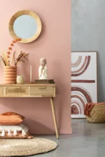 Een houten consoletafel staat tegen een roze muur, bedekt met decor. Daarboven hangt een ronde spiegel met rieten frame. Het Vloerkleed Rond Gevlochten Zeegras Naturel, kussens en een gevlochten mand sieren de vloer vlakbij.