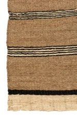 Het Vloerkleed Gevlochten Zeegras-Palmblad Naturel/Zwart is een geweven vloerkleed van natuurlijke vezels met horizontale zwarte, beige en donkerbruine strepen, gedeeltelijk met rafelige randen aan de linkerkant.