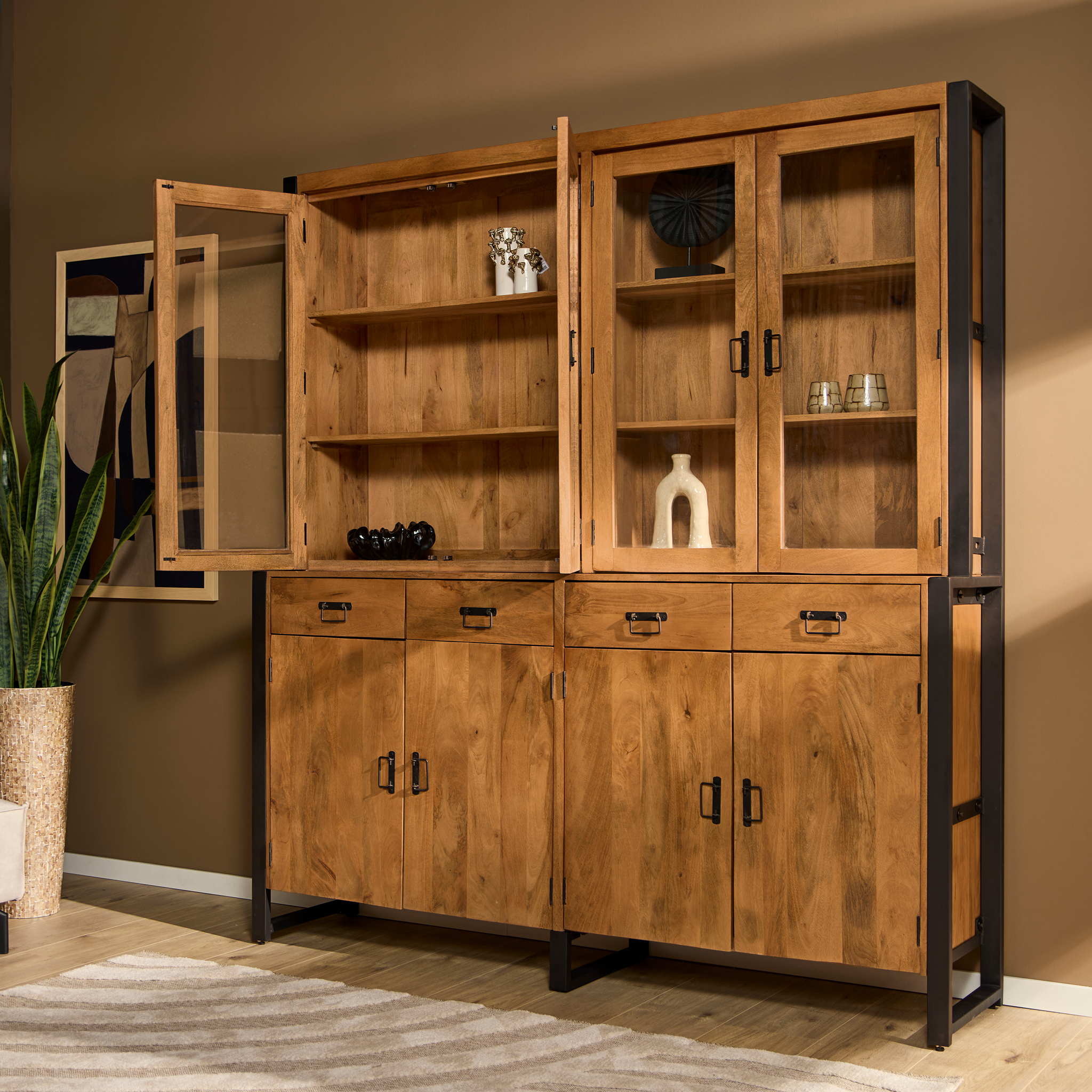 De Vitrinekast Boaz XXL, een grote houten kast met zwart metalen accenten, staat tegen een bruine muur. De open glazen deuren onthullen gedecoreerde planken. Een potplant en een ingelijst kunstwerk maken de stijlvolle omgeving compleet.