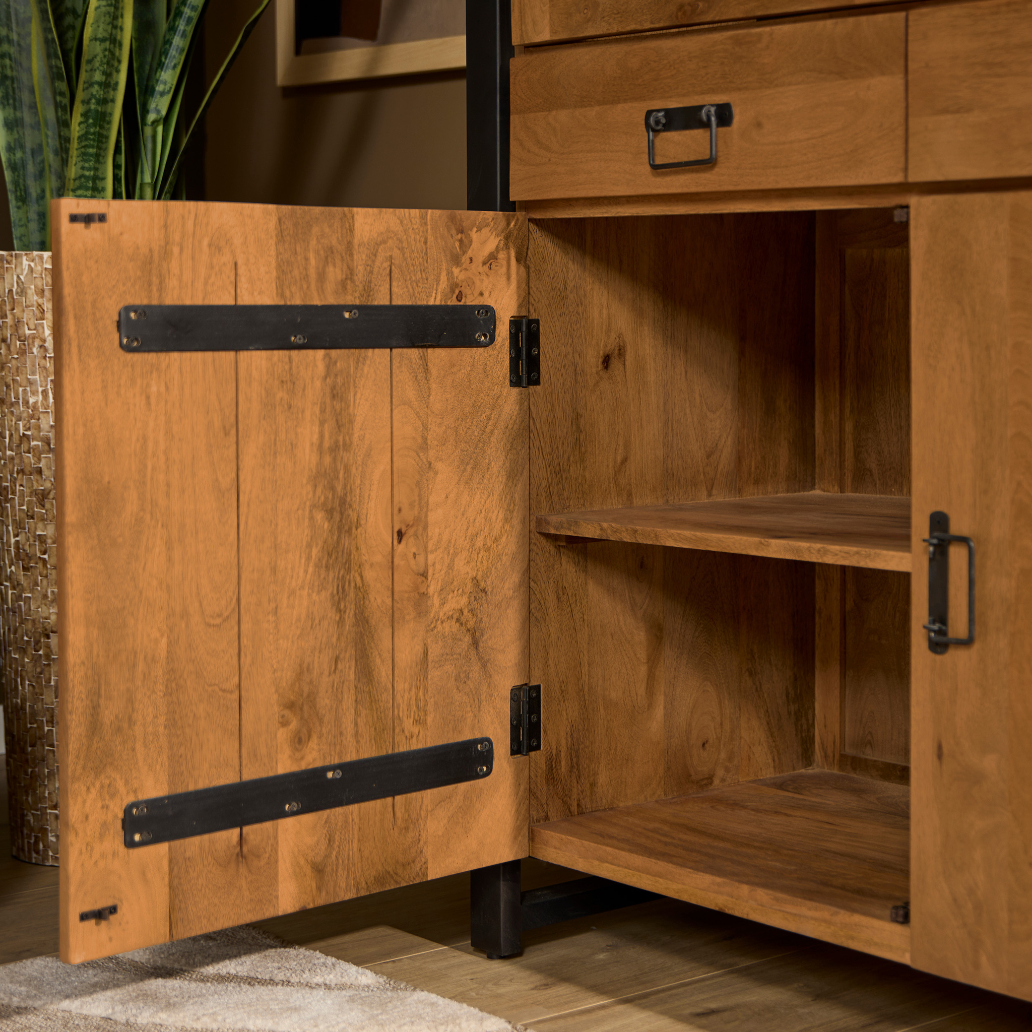 De Vitrinekast Boaz 115 cm, een houten kast met twee planken en zwart metalen scharnieren en handgrepen, wordt tentoongesteld met de deur open. Vlakbij zijn ook een plant in een pot met structuur en een deel van een beige vloerkleed te zien.