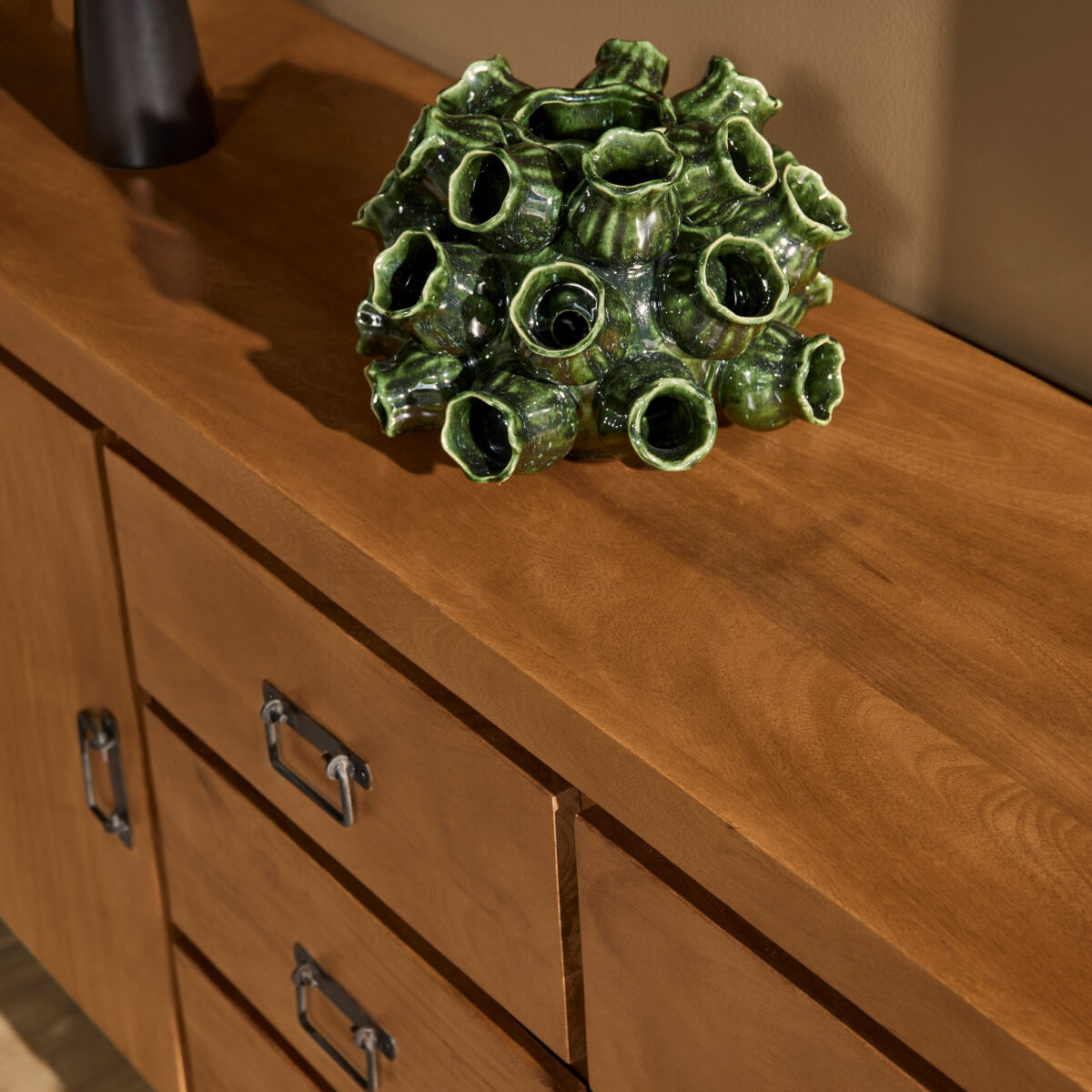 Een groene keramische sculptuur met verschillende holle buizen rust op de Dressoir Boaz 200 cm, een houten dressoir met metalen handgrepen, tegen een beige muur.