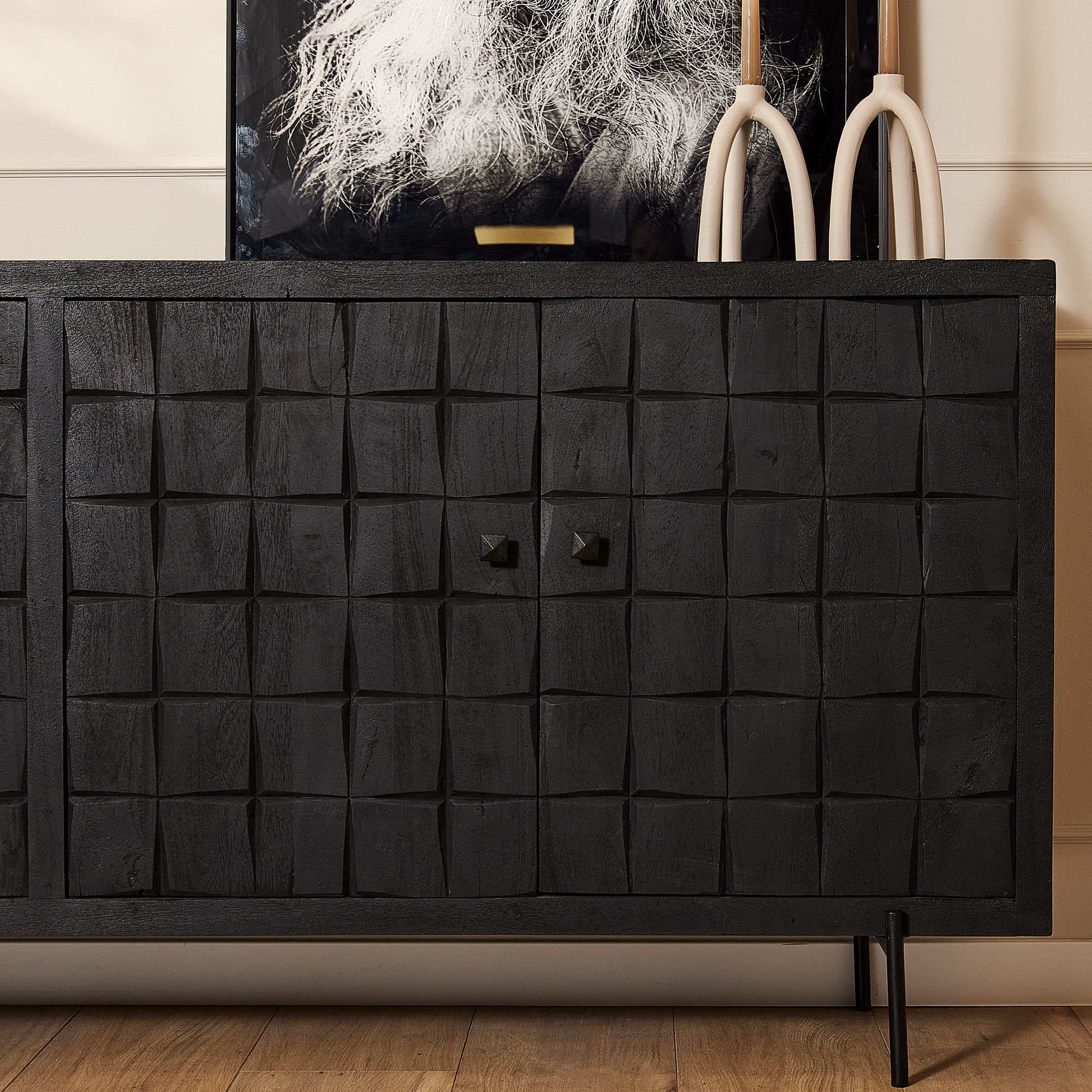 Een close-up van de Dressoir Brandy Black 220 cm toont het moderne gestructureerde oppervlak met vierkante patronen tegen een witte muur. Bovenop staan een zwart-wit kunstwerk en twee hoge decorstukken. De vloer is van hout.