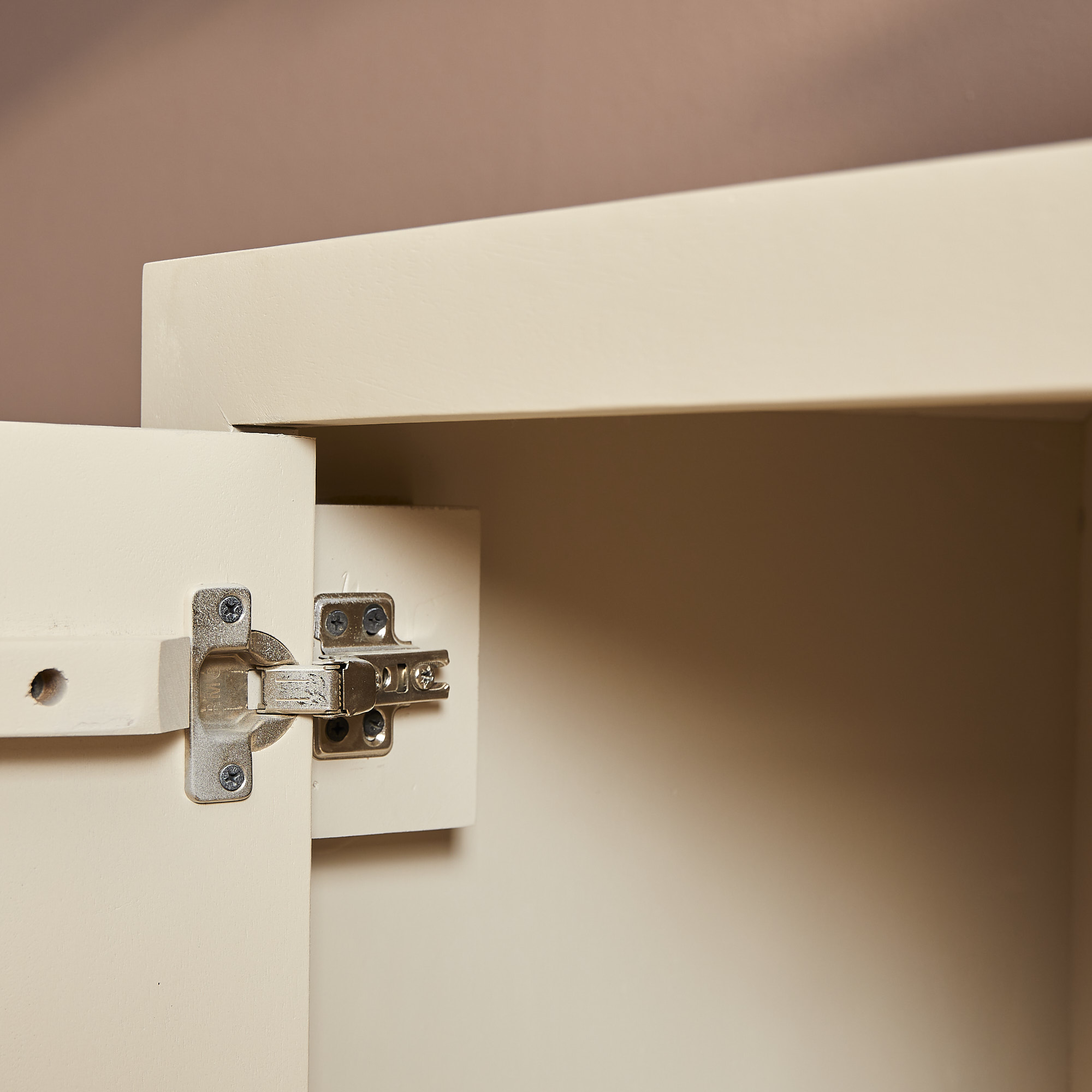 Close-up van een metalen scharnier aan de binnenhoek van een open Dressoir Brussel 3 deurs 150 cm crèmekleurige kastdeur tegen een zachtbruine muur.