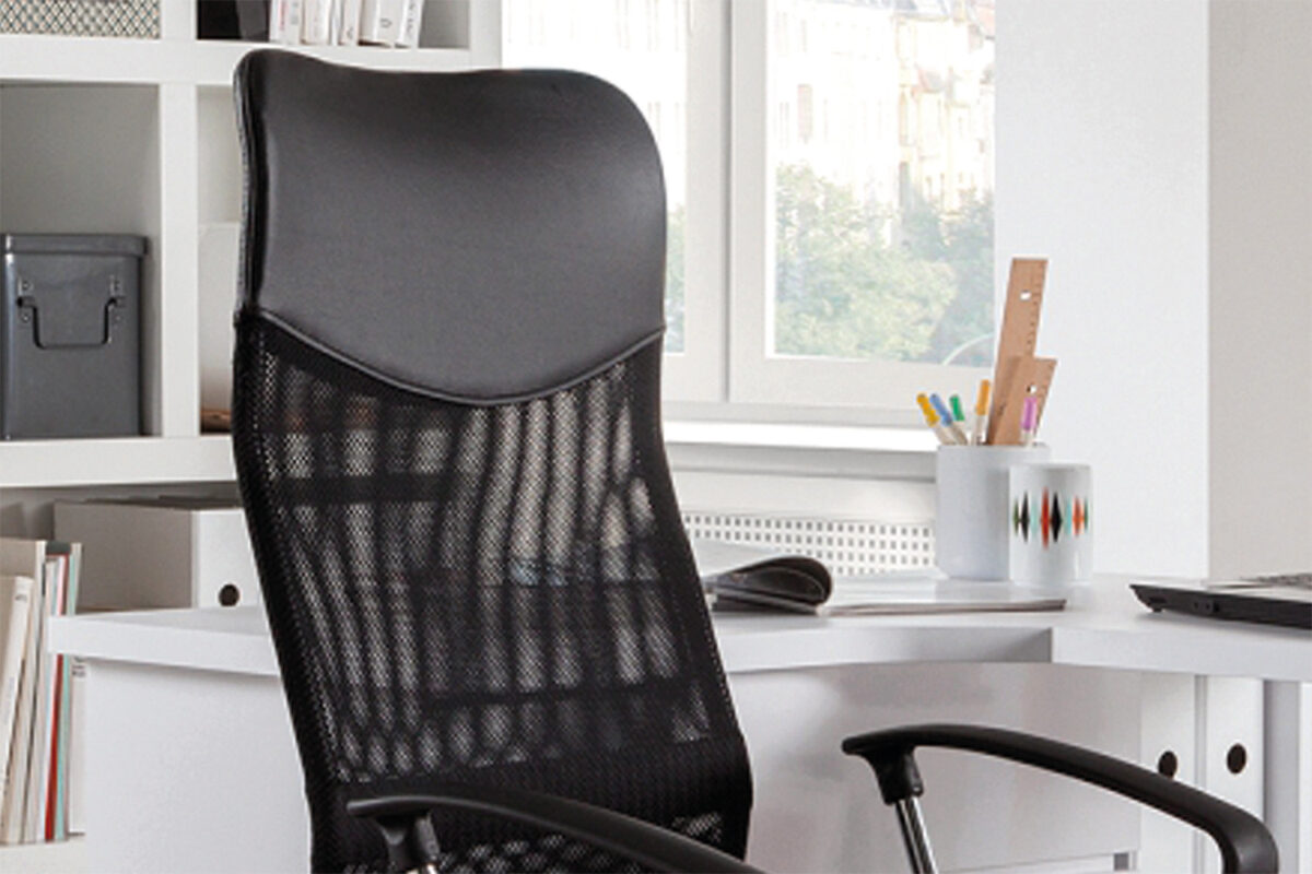 De Bureaustoel 'Max' Mesh Zwart (1c) staat voor een wit bureau in een lichte kamer met planken, boeken en een raam op de achtergrond.