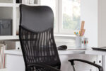 De Bureaustoel 'Max' Mesh Zwart (1c) staat voor een wit bureau in een lichte kamer met planken, boeken en een raam op de achtergrond.