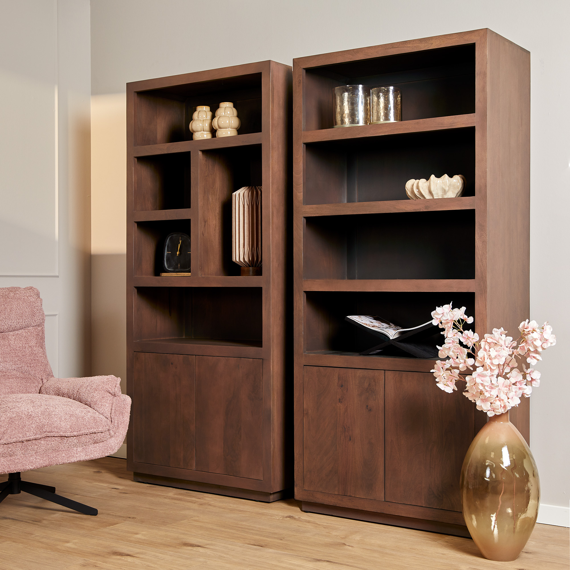 Twee Boekenkast Brussel Walnut | 90 cm boekenplanken met vazen, schalen en boeken staan naast een grote bruine vaas met roze bloemen. Een roze gestoffeerde fauteuil is gedeeltelijk zichtbaar aan de linkerkant, alles opgesteld op een houten vloer.